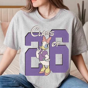 May include: A gray t-shirt featuring a purple graphic with the text "Class of '26" and a cartoon of Daisy Duck. Daisy Duck is wearing a purple dress and a pink bow. The shirt is a casual style.