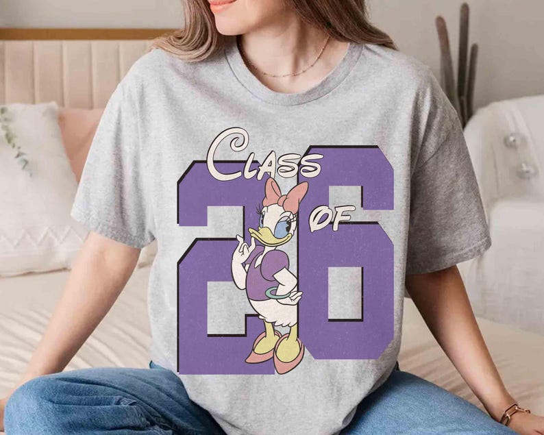 May include: A gray t-shirt featuring a purple graphic with the text "Class of '26" and a cartoon of Daisy Duck. Daisy Duck is wearing a purple dress and a pink bow. The shirt is a casual style.