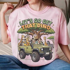 May include: Pink t-shirt with a graphic featuring cartoon characters in a green jeep with the text "LET'S GO GET THAT DINO". The jeep is surrounded by cartoon animals and a large tree.