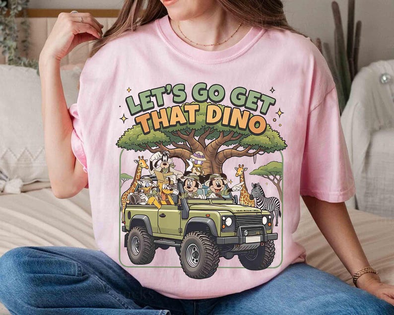 May include: Pink t-shirt with a graphic featuring cartoon characters in a green jeep with the text "LET'S GO GET THAT DINO". The jeep is surrounded by cartoon animals and a large tree.