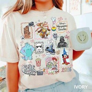 May include: Ivory-colored t-shirt featuring colorful Disney characters and icons, including Sully, Darth Vader, and Mickey Mouse. The shirt has the text "Disney's Hollywood Studios" and the year "2026". A white mug with a smiley face is also visible.