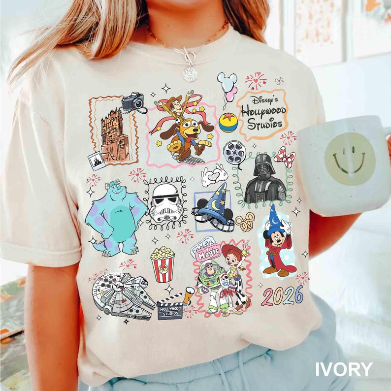 May include: Ivory-colored t-shirt featuring colorful Disney characters and icons, including Sully, Darth Vader, and Mickey Mouse. The shirt has the text "Disney's Hollywood Studios" and the year "2026". A white mug with a smiley face is also visible.