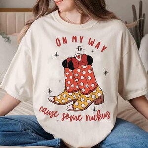Peut inclure: T-shirt blanc avec un motif de bottes rouges et jaunes &agrave; pois blancs. Le texte "On my way to cause some ruckus" est imprim&eacute; sur le t-shirt.