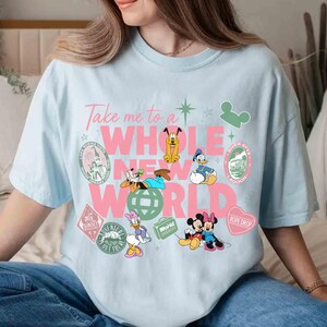 May include: Light blue t-shirt with the phrase "Take me to a WHOLE NEW WORLD" in pink and white. Features cartoon characters like Mickey and Minnie Mouse, Donald Duck, and Goofy, with travel-themed graphics.