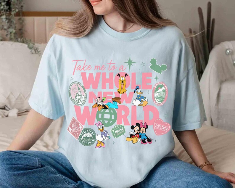 May include: Light blue t-shirt with the phrase "Take me to a WHOLE NEW WORLD" in pink and white. Features cartoon characters like Mickey and Minnie Mouse, Donald Duck, and Goofy, with travel-themed graphics.