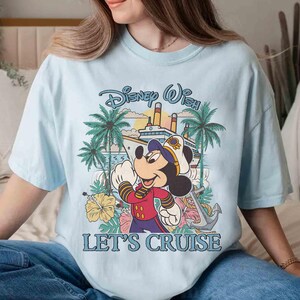 May include: Light blue t-shirt featuring Mickey Mouse in a captain's uniform with a cruise ship and palm trees. The text "Disney Wish" and "Let's Cruise" are displayed in a playful font. The shirt is a relaxed fit.