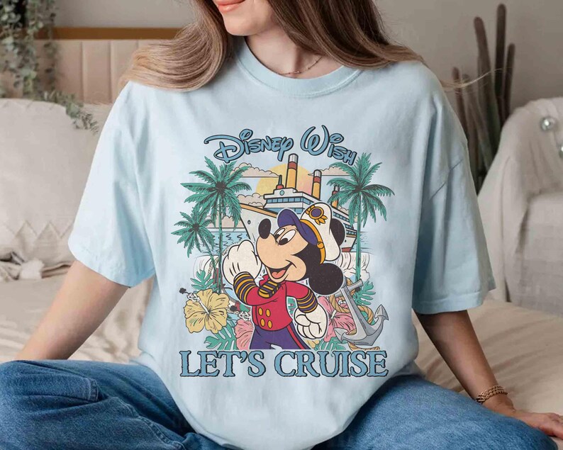 May include: Light blue t-shirt featuring Mickey Mouse in a captain's uniform with a cruise ship and palm trees. The text "Disney Wish" and "Let's Cruise" are displayed in a playful font. The shirt is a relaxed fit.