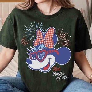 Op de afbeelding: Donkergroen t-shirt met een cartoon Minnie Mouse-ontwerp in rood, wit en blauw. Minnie Mouse draagt een hartvormige bril en een strik met een rood-wit geblokt patroon. De tekst "Red White & Cute" staat eronder.