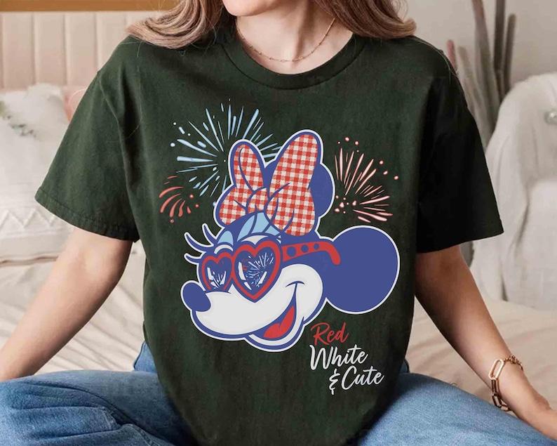 Op de afbeelding: Donkergroen t-shirt met een cartoon Minnie Mouse-ontwerp in rood, wit en blauw. Minnie Mouse draagt een hartvormige bril en een strik met een rood-wit geblokt patroon. De tekst "Red White & Cute" staat eronder.