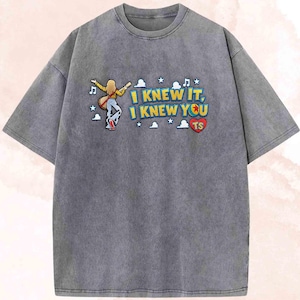 May include: A charcoal grey acid-wash t-shirt featuring a graphic of a person with long blonde hair playing a guitar. The text reads I KNEW IT, I KNEW YOU with a red heart containing the letters TS, surrounded by small white clouds, stars, and musical notes.