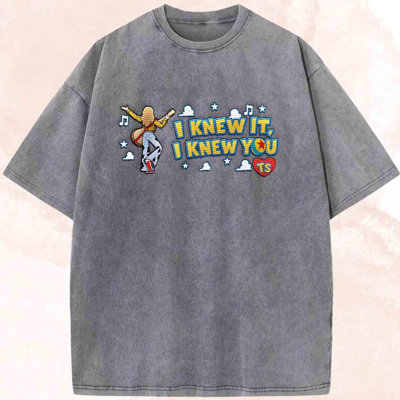 May include: A charcoal grey acid-wash t-shirt featuring a graphic of a person with long blonde hair playing a guitar. The text reads I KNEW IT, I KNEW YOU with a red heart containing the letters TS, surrounded by small white clouds, stars, and musical notes.
