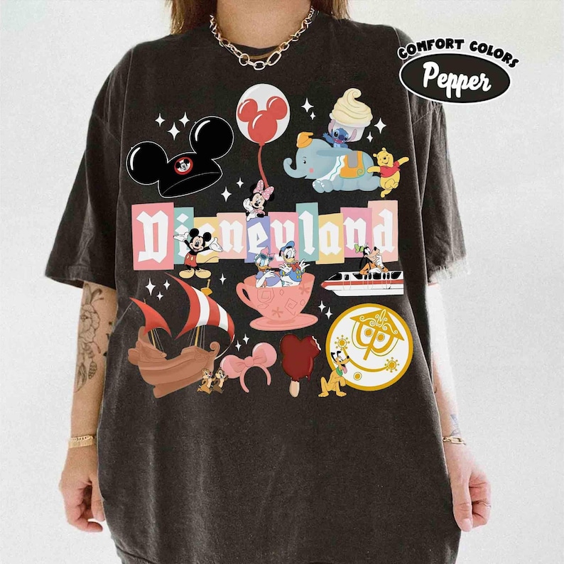May include: Dark gray t-shirt featuring a colorful Disneyland graphic. The design includes Mickey Mouse ears, various Disney characters, and the Disneyland logo. The shirt has a vintage, distressed look with the words "Comfort Colors Pepper" printed on the upper right.