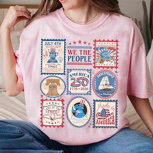 以下が含まれることがあります： 様々な愛国的な切手デザインが施された、薄いピンク色のTシャツ。自由の鐘、船、アメリカ国旗の画像が切手に描かれており、「July 4th」や「Made in America」などのテキストが含まれています。
