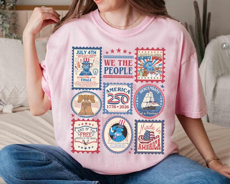 以下が含まれることがあります： 様々な愛国的な切手デザインが施された、薄いピンク色のTシャツ。自由の鐘、船、アメリカ国旗の画像が切手に描かれており、「July 4th」や「Made in America」などのテキストが含まれています。