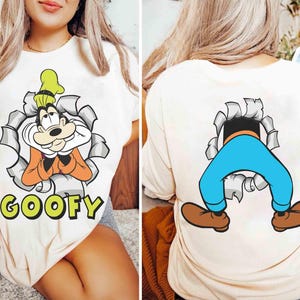 Może przedstawiać: Kremowa koszulka z kreskówkowym wizerunkiem Goofy'ego przebijającego się przez efekt podartego papieru, z napisem "GOOFY" w zielonych literach. Tył koszulki przedstawia nogi i stopy Goofy'ego. Zabawny, zabawny design.