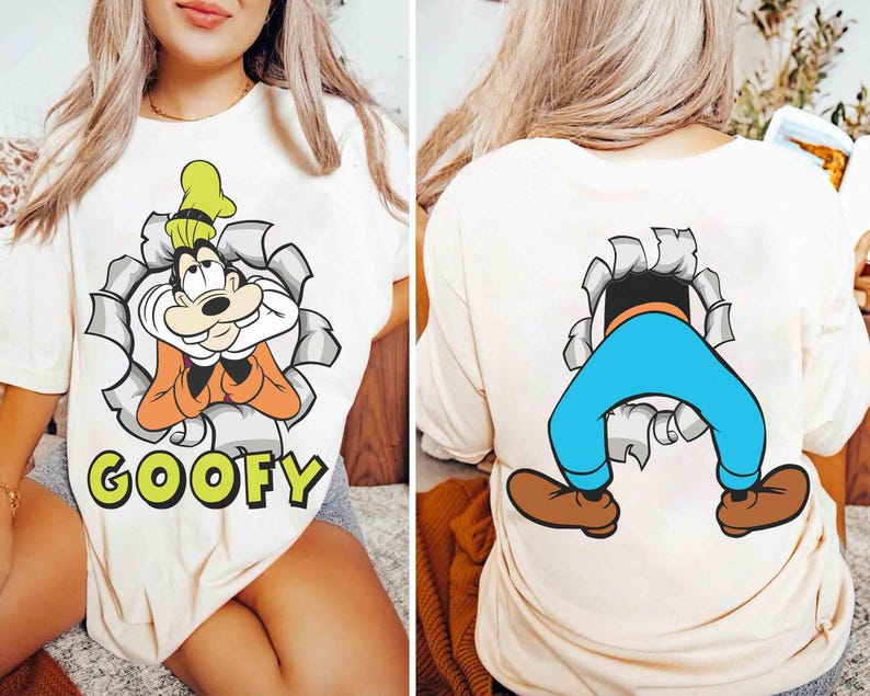 May include: Cream-colored t-shirt featuring a cartoon image of Goofy bursting through a torn paper effect, with the name "GOOFY" in green letters. The back of the shirt shows Goofy's legs and feet. A fun, playful design.