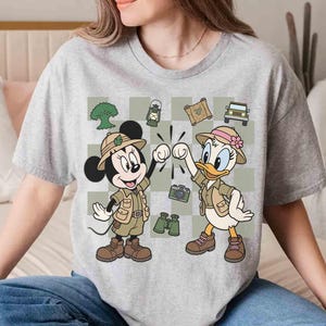 May include: Gray t-shirt featuring Mickey Mouse and Daisy Duck in safari outfits, giving a fist bump. The design includes a checkered background with safari-themed illustrations like a jeep, map, and binoculars.