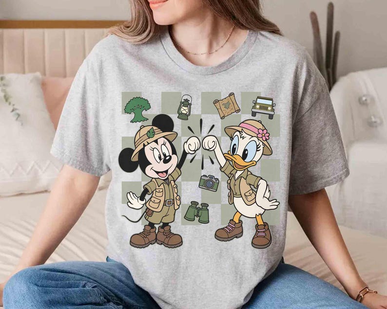 May include: Gray t-shirt featuring Mickey Mouse and Daisy Duck in safari outfits, giving a fist bump. The design includes a checkered background with safari-themed illustrations like a jeep, map, and binoculars.