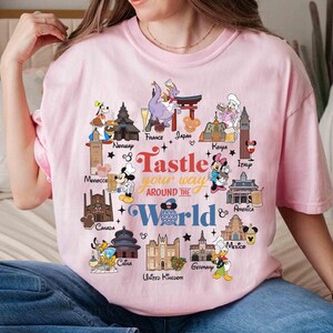 May include: A light pink t-shirt with a colorful graphic design. The design features cartoon characters and landmarks from around the world, with the text "Taste Your Way Around The World".