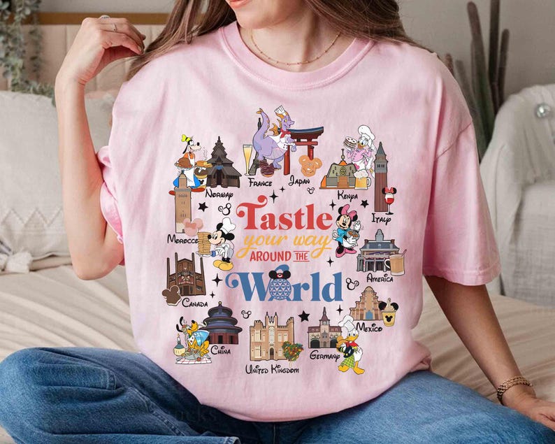 May include: A light pink t-shirt with a colorful graphic design. The design features cartoon characters and landmarks from around the world, with the text "Taste Your Way Around The World".