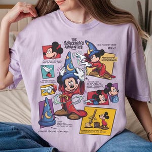 May include: A light purple t-shirt featuring a graphic of Mickey Mouse as the Sorcerer's Apprentice. The design includes multiple panels with illustrations and text from the 1940 Disney film, Fantasia.