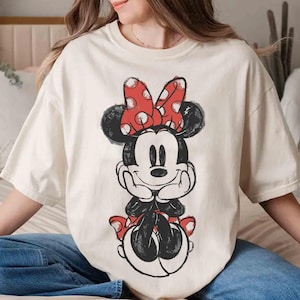 May include: A cream-coloured t-shirt with a distressed Minnie Mouse graphic. Minnie is in black and white with a red polka-dot bow. The shirt has short sleeves and a relaxed fit, suitable for casual wear.