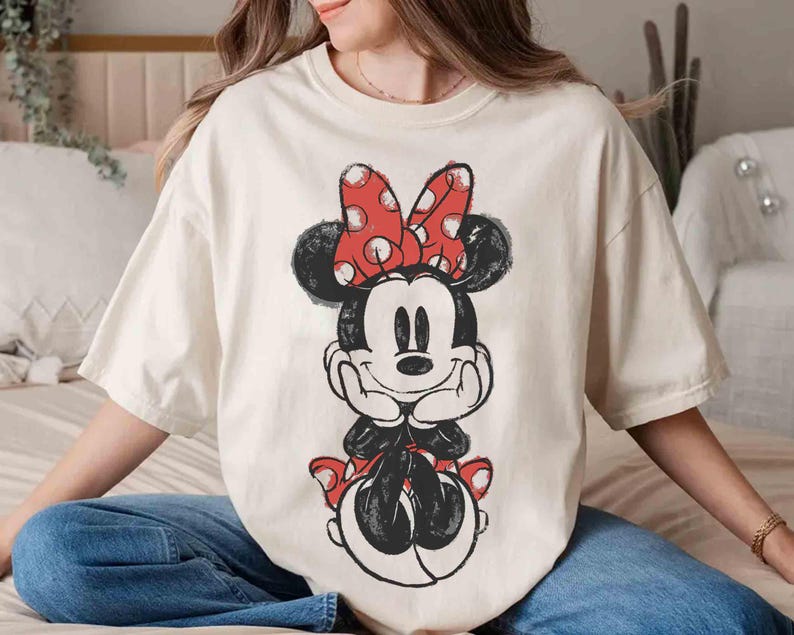 May include: A cream-coloured t-shirt with a distressed Minnie Mouse graphic. Minnie is in black and white with a red polka-dot bow. The shirt has short sleeves and a relaxed fit, suitable for casual wear.