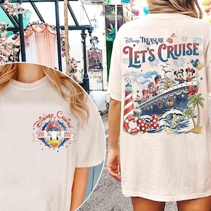May include: A cream-colored t-shirt featuring a small graphic of Donald Duck on the front left chest with the text Disney Cruise 2026. The back displays a large illustration of a cruise ship, Mickey and Minnie Mouse, a lighthouse, and Let's Cruise.