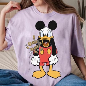 May include: A lilac t-shirt with a cartoon Donald Duck design. The character has a white face, black body, red shorts, and yellow feet. The shirt includes the text "QUACK!" with star graphics.