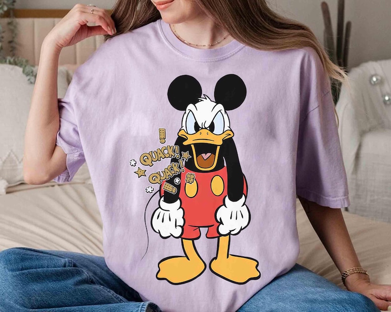 May include: A lilac t-shirt with a cartoon Donald Duck design. The character has a white face, black body, red shorts, and yellow feet. The shirt includes the text "QUACK!" with star graphics.
