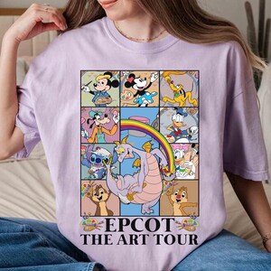 May include: A light purple t-shirt featuring a colorful graphic of Disney characters in a grid pattern. The graphic includes Mickey Mouse, Minnie Mouse, Donald Duck, and Stitch, with the text "EPCOT THE ART TOUR" at the bottom.