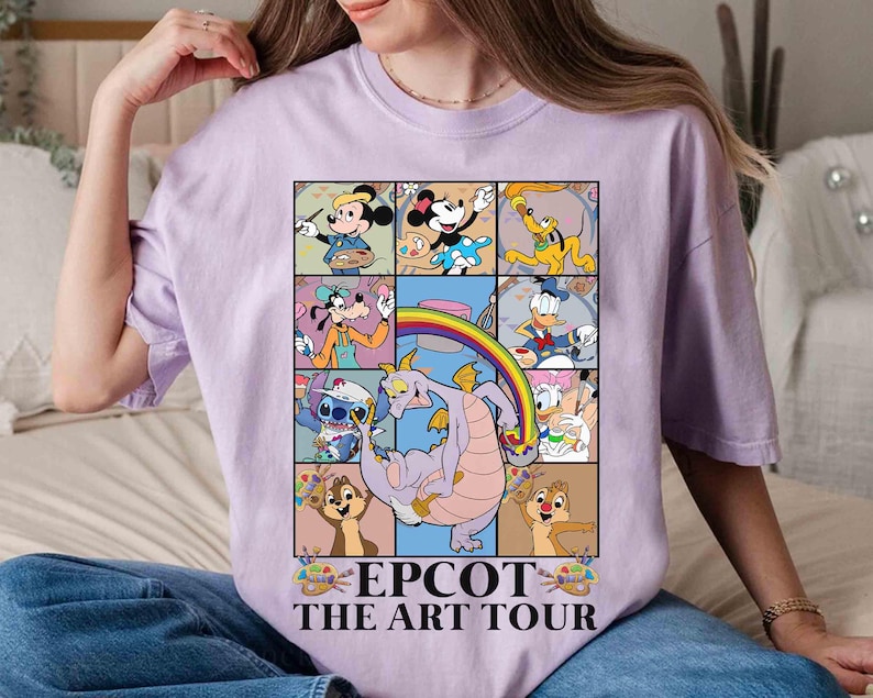 May include: A light purple t-shirt featuring a colorful graphic of Disney characters in a grid pattern. The graphic includes Mickey Mouse, Minnie Mouse, Donald Duck, and Stitch, with the text "EPCOT THE ART TOUR" at the bottom.