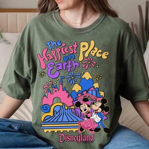 K&ouml;nnte beinhalten: Olivgr&uuml;nes T-Shirt mit einem farbenfrohen Aufdruck von Mickey und Minnie Mouse vor dem Disneyland-Schloss. Der Text "The Happiest Place on Earth" und "Disneyland Resort" ist ebenfalls auf dem Shirt zu sehen.