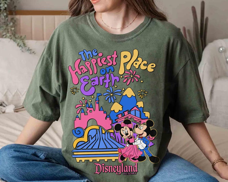 K&ouml;nnte beinhalten: Olivgr&uuml;nes T-Shirt mit einem farbenfrohen Aufdruck von Mickey und Minnie Mouse vor dem Disneyland-Schloss. Der Text "The Happiest Place on Earth" und "Disneyland Resort" ist ebenfalls auf dem Shirt zu sehen.