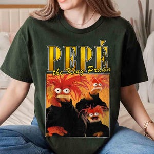 May include: A dark green t-shirt with a graphic print of three cartoon characters. The characters are red, orange, and yellow and have large eyes. The text "PEPE the King Prawn" is printed in yellow letters on the shirt.
