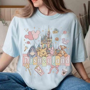 May include: Light blue t-shirt featuring a Disneyland graphic. The design includes a castle, balloons, and various Disney characters such as Winnie the Pooh, Piglet, Tigger, and Eeyore. Additional elements include popcorn, a bow, and a Mickey Mouse-shaped drink.