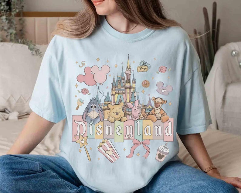 May include: Light blue t-shirt featuring a Disneyland graphic. The design includes a castle, balloons, and various Disney characters such as Winnie the Pooh, Piglet, Tigger, and Eeyore. Additional elements include popcorn, a bow, and a Mickey Mouse-shaped drink.