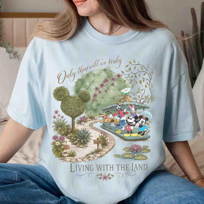 May include: Light blue t-shirt featuring a whimsical garden scene with Disney characters. The design includes the text "Only then will we truly" and "Living with the Land". The graphic showcases Mickey and Minnie Mouse, and other characters.