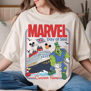 May include: Cream-colored t-shirt featuring a graphic of a cruise ship with the text "MARVEL Day at Sea". The design includes a cartoon character and Mickey Mouse icons. The words "Custom Name" are also visible.