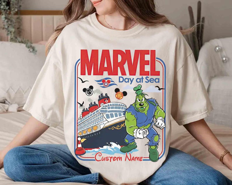 May include: Cream-colored t-shirt featuring a graphic of a cruise ship with the text "MARVEL Day at Sea". The design includes a cartoon character and Mickey Mouse icons. The words "Custom Name" are also visible.