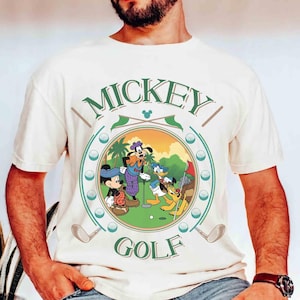 May include: Cream-colored t-shirt featuring a circular graphic of Mickey Mouse, Goofy, Donald Duck, and Pluto playing golf. The graphic includes the words "MICKEY" and "GOLF" in green, with golf clubs and golf balls around the circle.