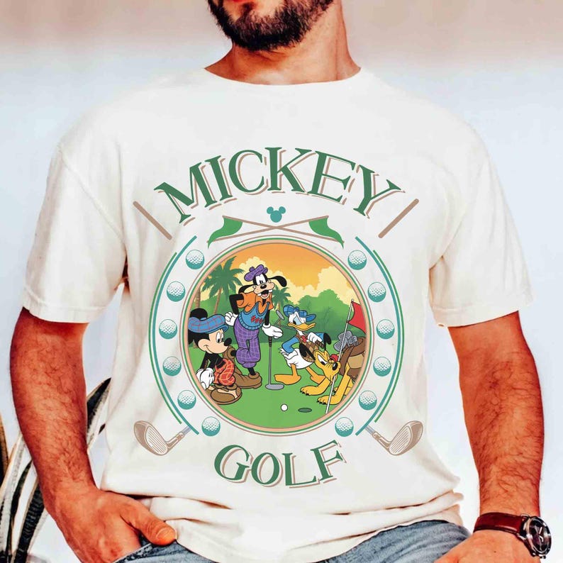 May include: Cream-colored t-shirt featuring a circular graphic of Mickey Mouse, Goofy, Donald Duck, and Pluto playing golf. The graphic includes the words "MICKEY" and "GOLF" in green, with golf clubs and golf balls around the circle.
