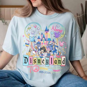May include: Light blue t-shirt featuring a colorful Disneyland design. The design includes the Disneyland logo, a castle, and various Disney characters. Text on the shirt reads "Disneyland 70 Years Magic 1955-2025" and "Name".