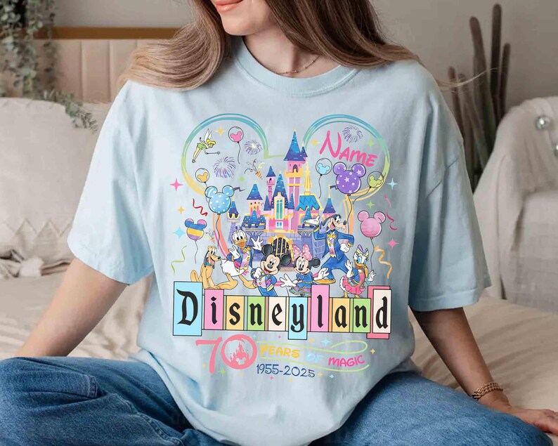 May include: Light blue t-shirt featuring a colorful Disneyland design. The design includes the Disneyland logo, a castle, and various Disney characters. Text on the shirt reads "Disneyland 70 Years Magic 1955-2025" and "Name".
