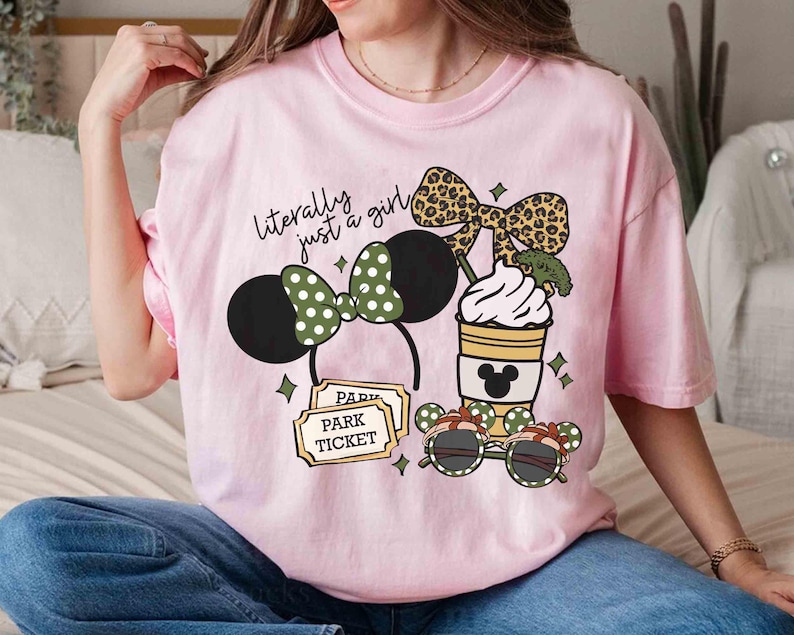 May include: Pink t-shirt with a graphic featuring a Minnie Mouse ear headband, a coffee cup, and sunglasses. The text "literally just a girl" is above the graphic. The design includes a park ticket.