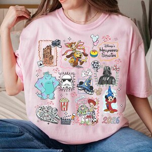 May include: A light pink t-shirt featuring various Disney characters and icons, including Sully, Darth Vader, and Mickey Mouse. The shirt has the text "Disney's Hollywood Studios" and the year "2026".