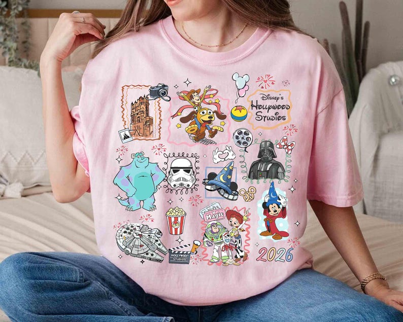 May include: A light pink t-shirt featuring various Disney characters and icons, including Sully, Darth Vader, and Mickey Mouse. The shirt has the text "Disney's Hollywood Studios" and the year "2026".