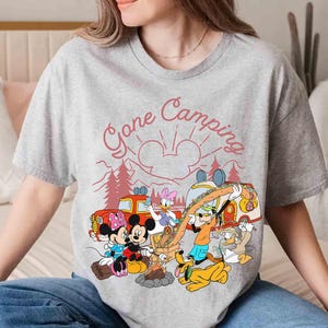Pu&ograve; includere: T-shirt grigia con grafica a tema campeggio con Topolino, Minnie, Paperino, Paperina, Pippo e Pluto. La grafica include il testo "Gone Camping" in rosso, con un design di sole e montagne.