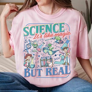 Pu&ograve; includere: T-shirt rosa chiaro con una grafica colorata che recita "SCIENCE It's like magic BUT REAL." La grafica presenta personaggi dei cartoni animati e illustrazioni scientifiche, tra cui beute, formule e un filamento di DNA.