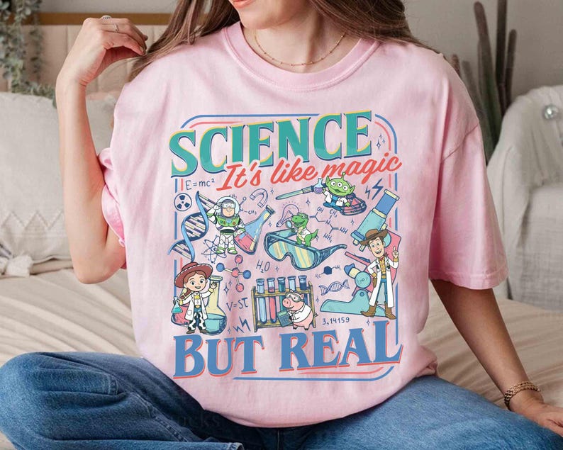 Pu&ograve; includere: T-shirt rosa chiaro con una grafica colorata che recita "SCIENCE It's like magic BUT REAL." La grafica presenta personaggi dei cartoni animati e illustrazioni scientifiche, tra cui beute, formule e un filamento di DNA.